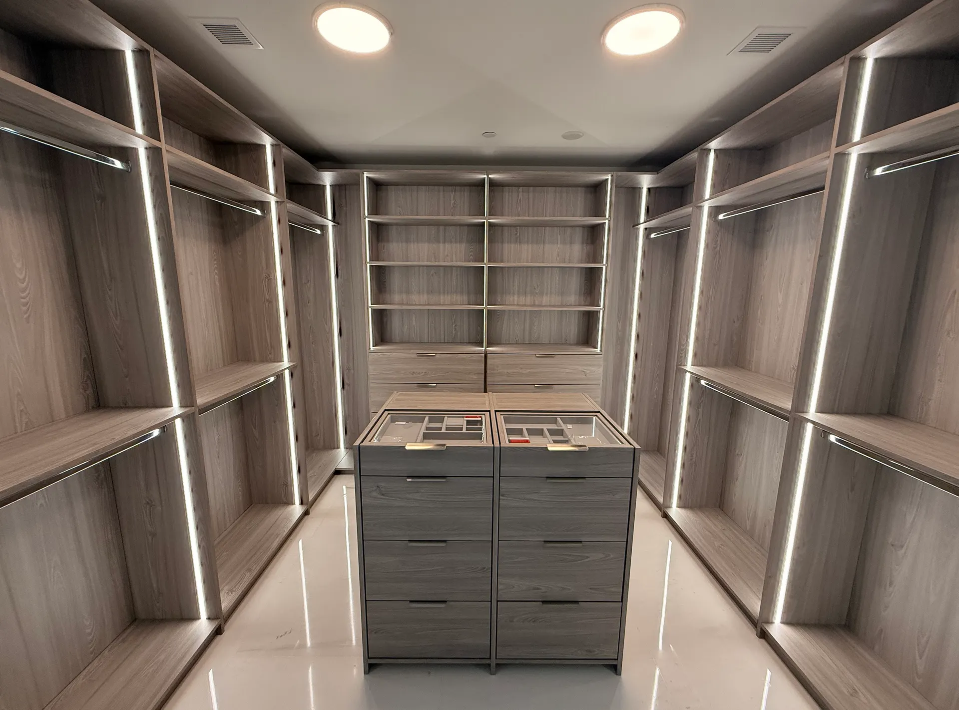 Finished walk-in closet in light grey wood, with LED-lit shelving on both walls and a central island of drawers with glass-topped jewellery trays