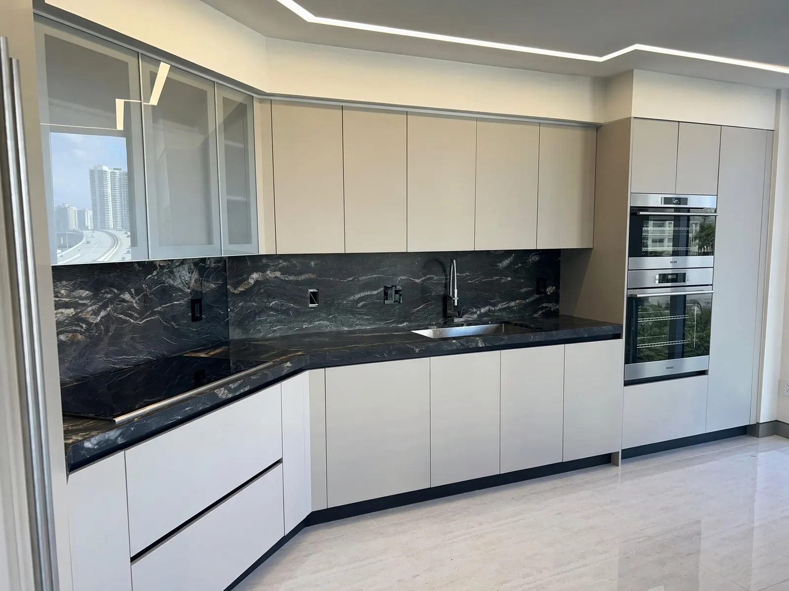 L-shaped kitchen with handleless greige cabinets, glass-front uppers, a dark veined marble counter and backsplash, a double wall oven and a city view