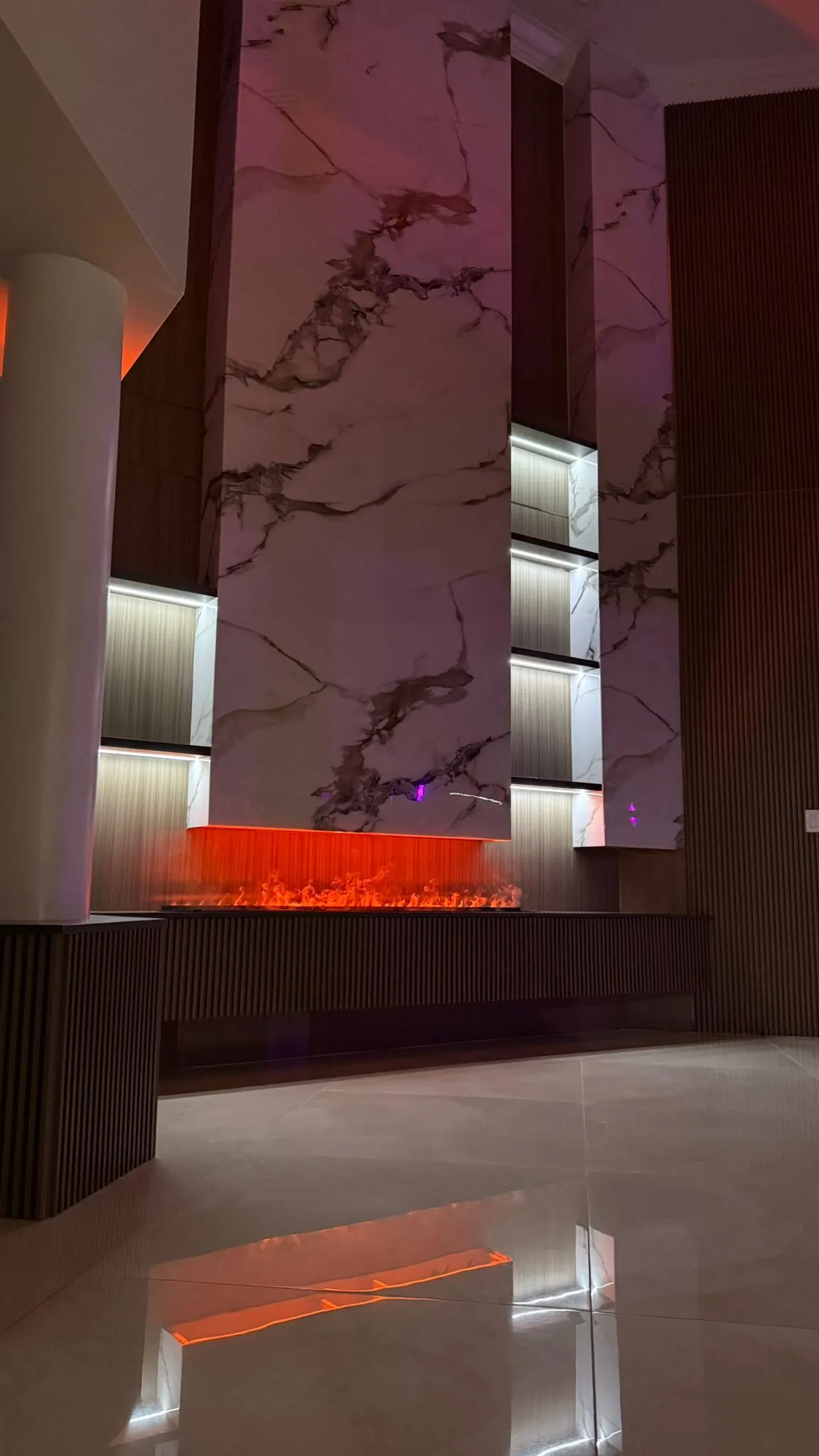 Double-height feature wall in marble-look panels and fluted wood at night, with the linear fireplace lit under the marble and the niches glowing