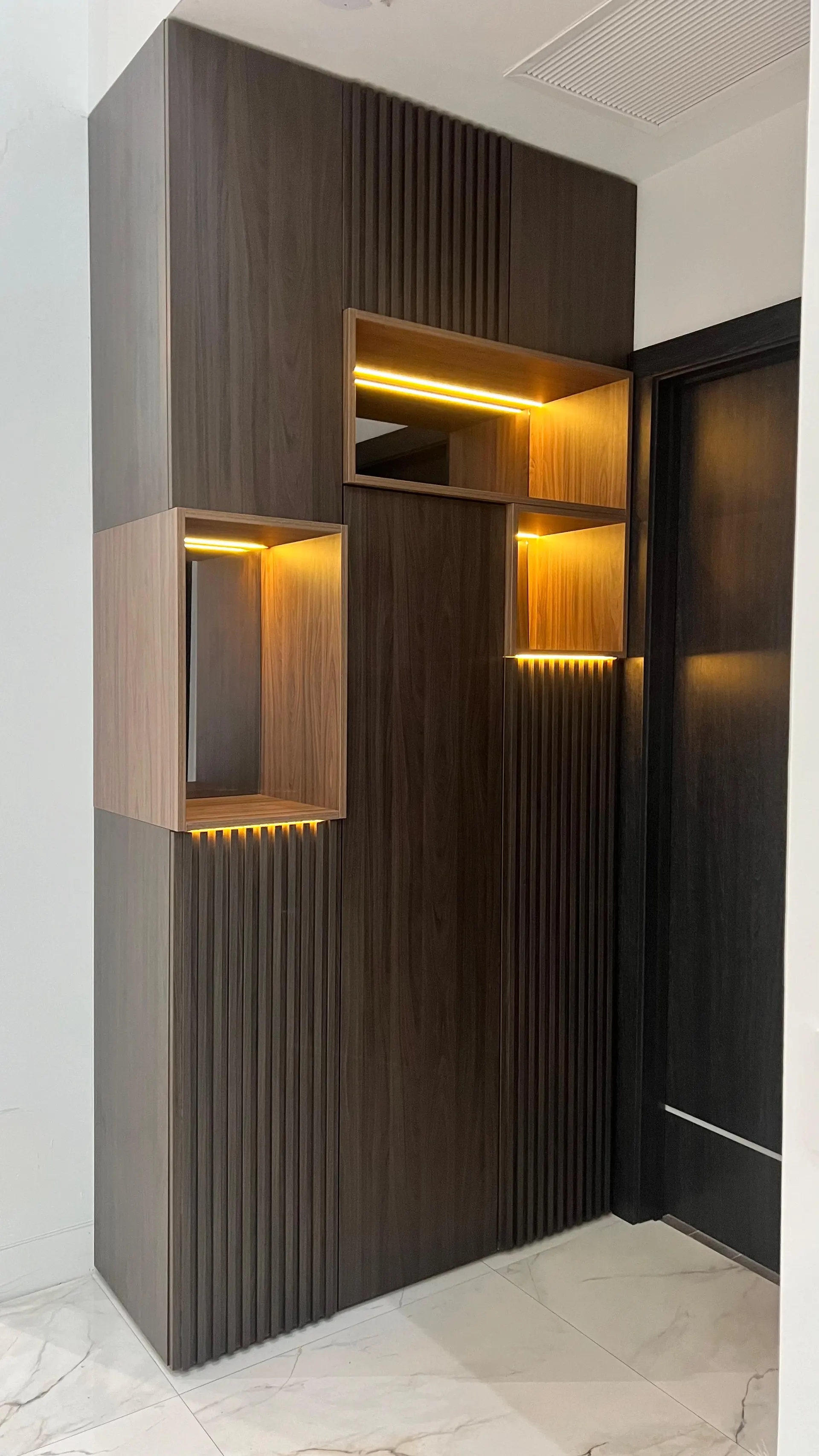 Floor-to-ceiling walnut unit with fluted panels and three lit display niches in lighter oak